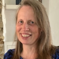 A woman with long light brown hair smiling, wearing a blue patterned top, indoors.