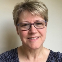 Smiling woman with short, fair hair and glasses, wearing a patterned top, in front of a plain background.