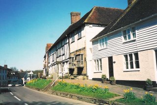 Old half-timbered houses and white cottages line a village street on a sunny day with daffodils in flower.