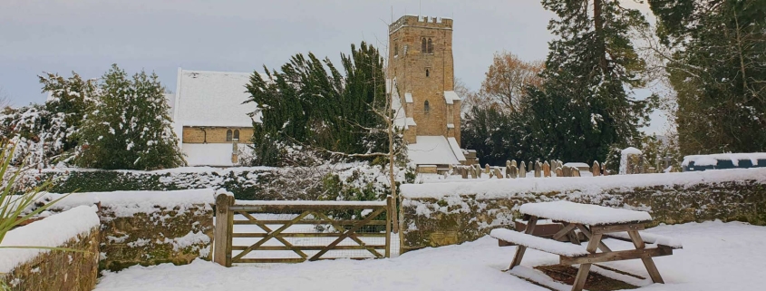 A snow-covered picnic table and gate with a church and trees in the background