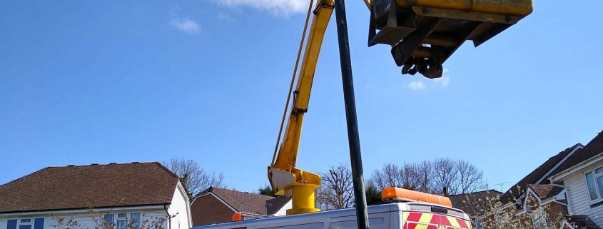 A worker in a lift repairs a streetlamp next to a utility van on a suburban road.