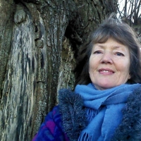 Woman in a blue scarf and coat standing beside a large tree, smiling at the camera.